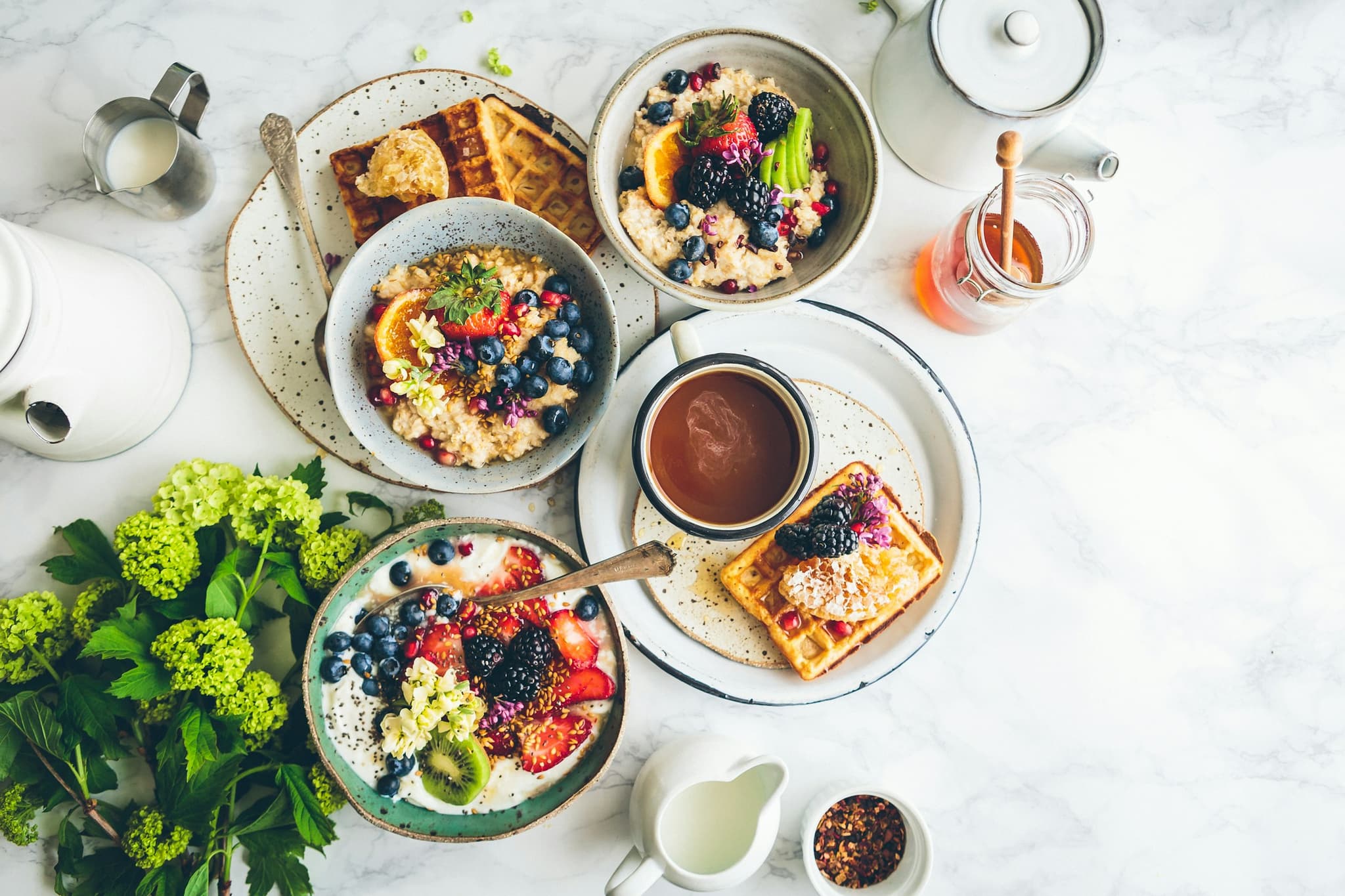 Five different high protein breakfasts arranged on a clean kitchen counter — overnight oats, a protein smoothie, eggs on toast, a yoghurt parfait, and a wrap in morning light