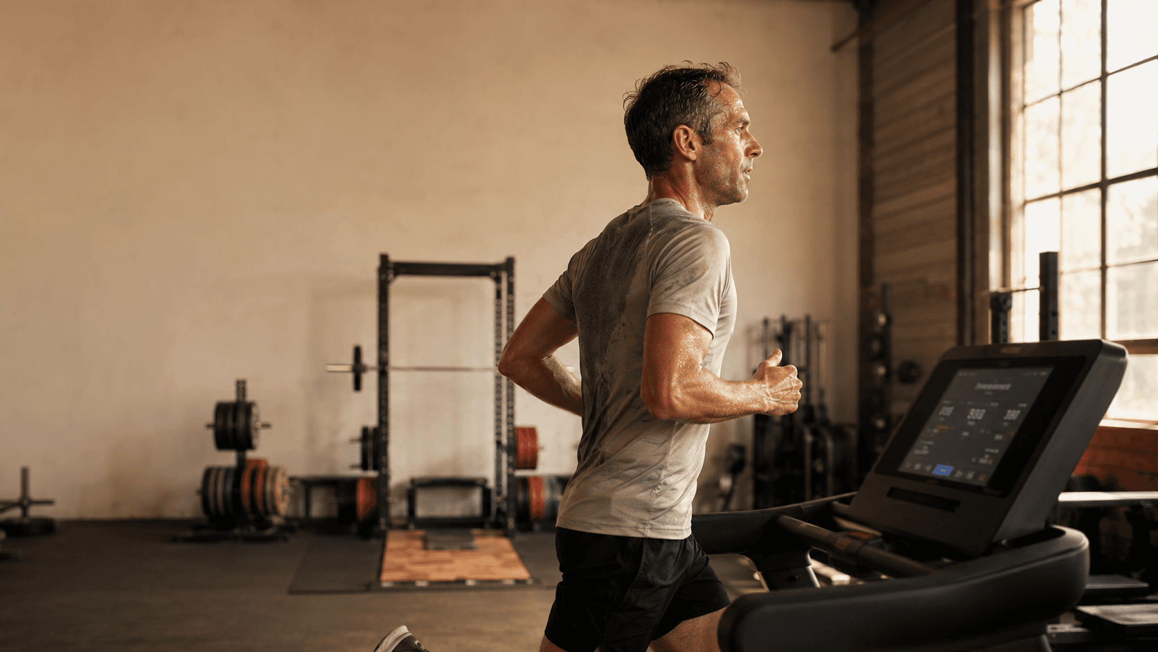 A lean, silver-haired man past 30 running on a treadmill in a sunlit gym with a barbell rack and dumbbell rack sitting unused in the background — capturing the cardio trap that keeps skinny fat men's weight stable while muscle quietly disappears year by year