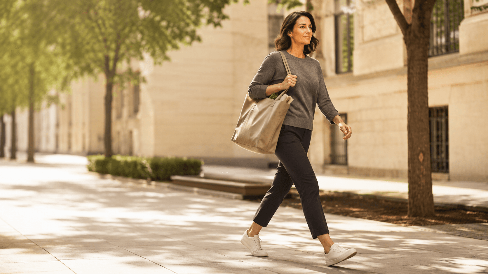 A woman walking briskly down a sunlit city street with a tote bag over her shoulder, mid stride and clearly moving with purpose — illustrating the NEAT message above that daily walking and unstructured movement, not extra gym cardio, is the biggest variable most women can control for fat loss
